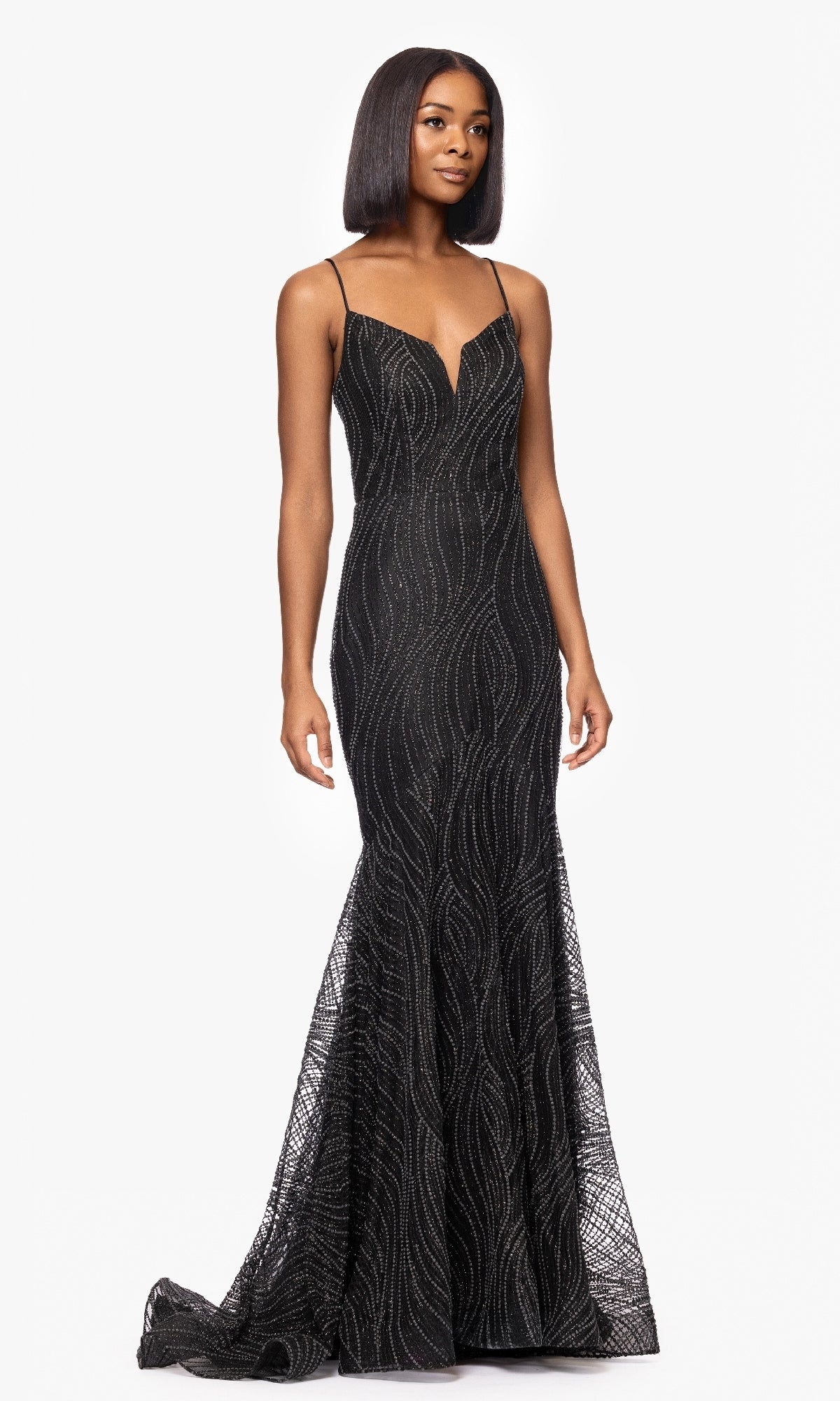 A woman poses against a white backdrop in the Blondie Nites 5491BN Long Black Glitter Prom Gownโ a fitted, floor-length dress with thin straps and a subtle wavy pattern, radiating timeless formal elegance.